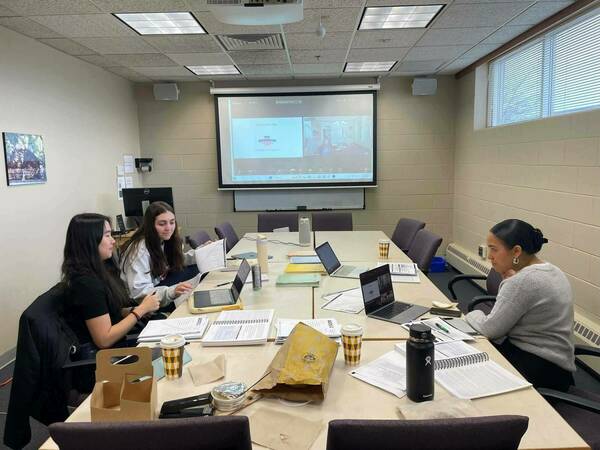 Three students participate in ADOS Training. They sit at a long table in a conference room, engaged with laptops and printed materials. A projector screen displays a presentation.